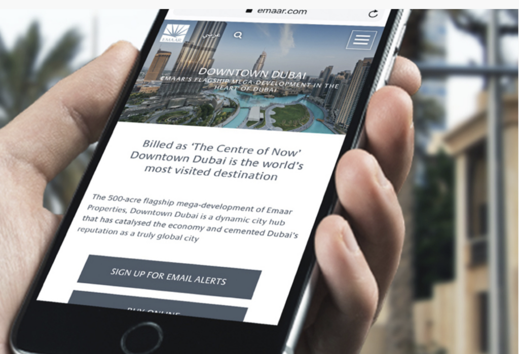 Hand holding a smartphone displaying a Downtown Dubai website or app interface with city imagery and text.