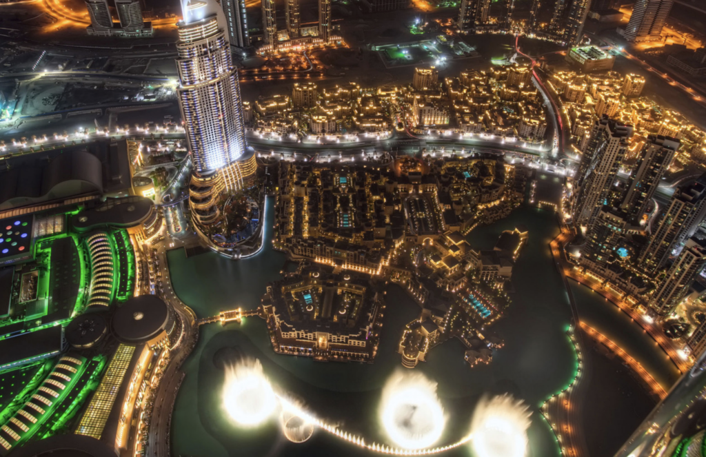 Nighttime aerial view of Downtown Dubai with illuminated buildings and the Dubai Fountain glowing below.