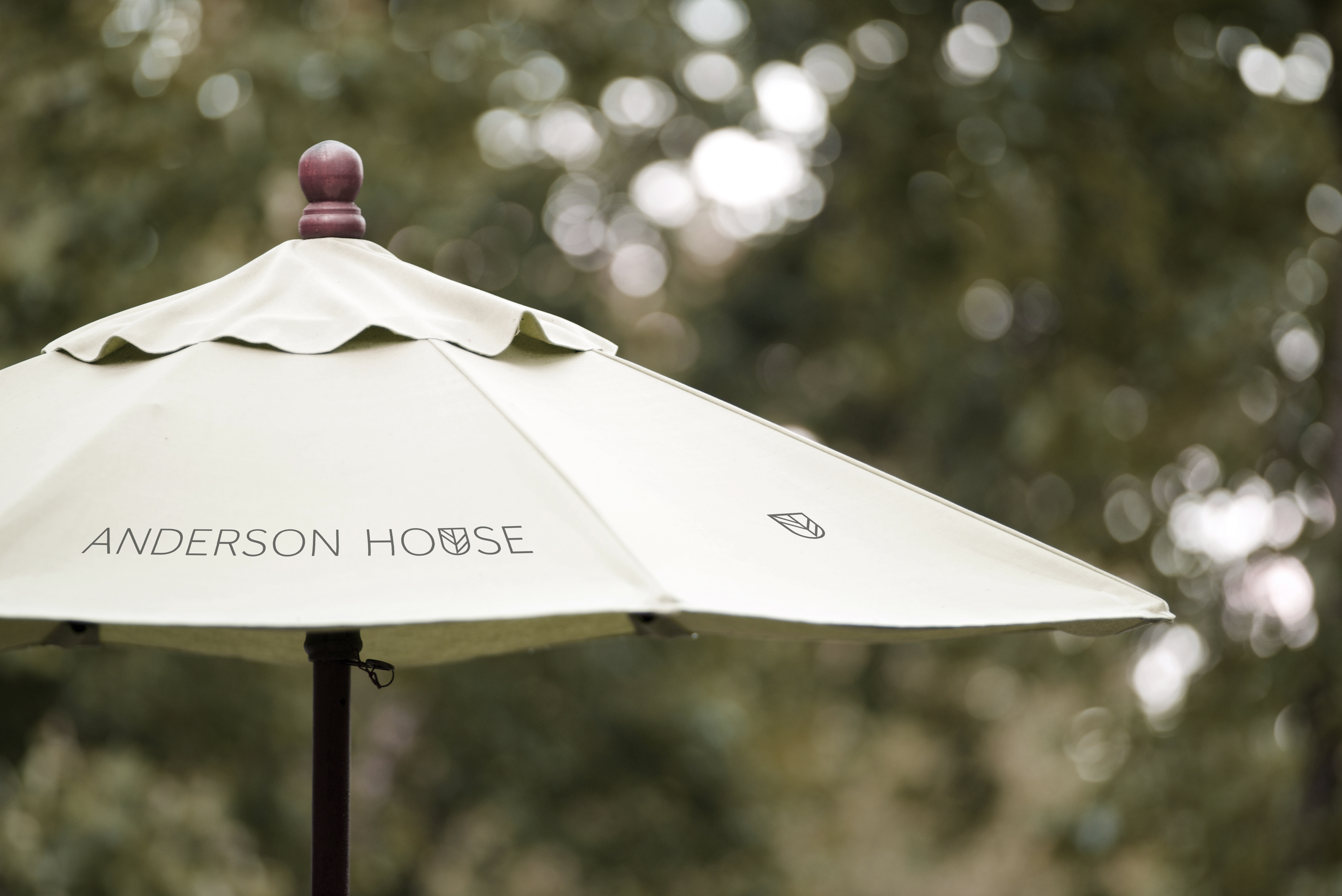 Outdoor parasol with “Anderson House” branding and logo, set against a softly blurred garden background.