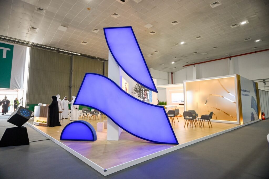 Exhibition booth featuring large curved illuminated blue sculptural elements in a spacious indoor event hall with seating and displays.