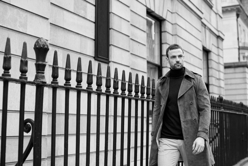 Black-and-white photo of a man in a coat walking past a wrought iron fence along a classic stone building façade.