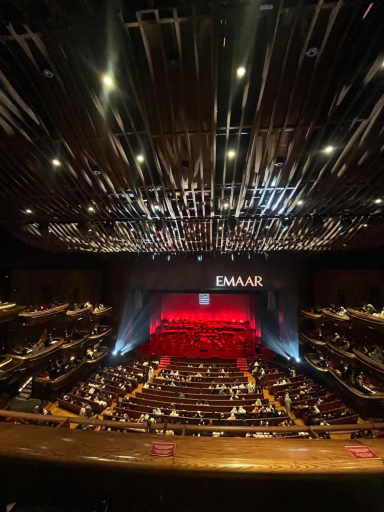 An interior view of a grand theater auditorium branded EMAAR, with tiered seating and a stage lit in red, hosting a live performance or event.